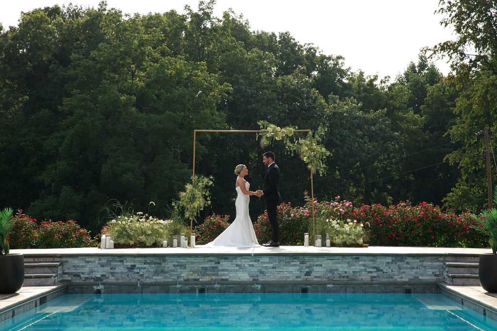 Newlyweds stand holding hands under their ceremony arbor on top of a pool surrounded by rose bushes washington dc wedding florist