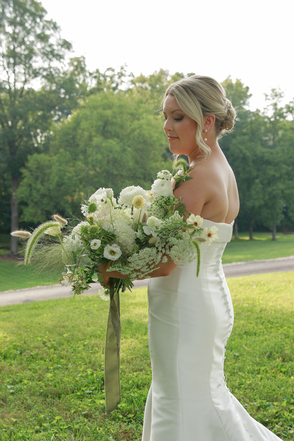 A bride in a white silk dress stands in a field gazing down on her white bouquet from a washington dc wedding florist