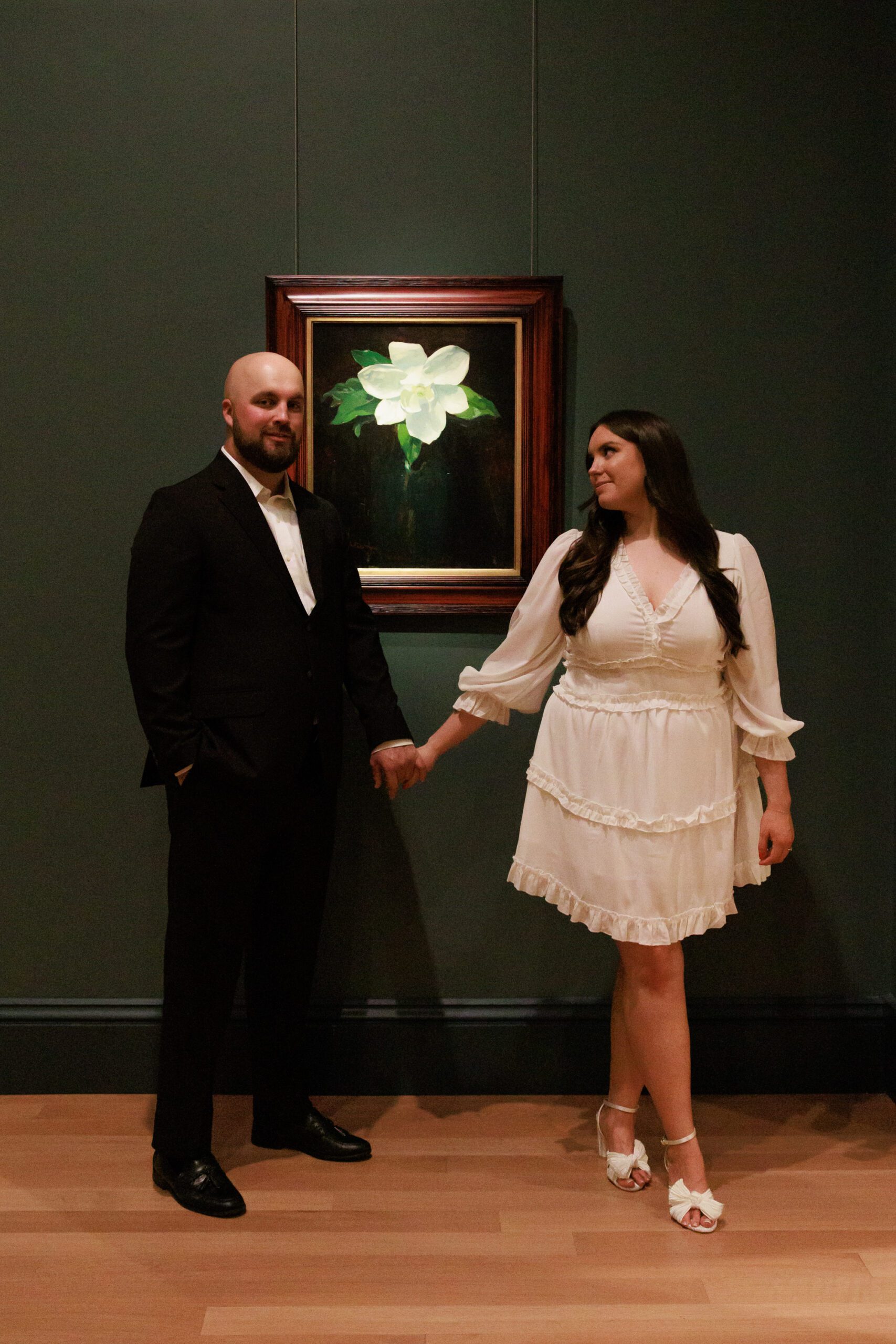 A couple holds hands for their engagement photos in front of a piece of art featuring a flower.