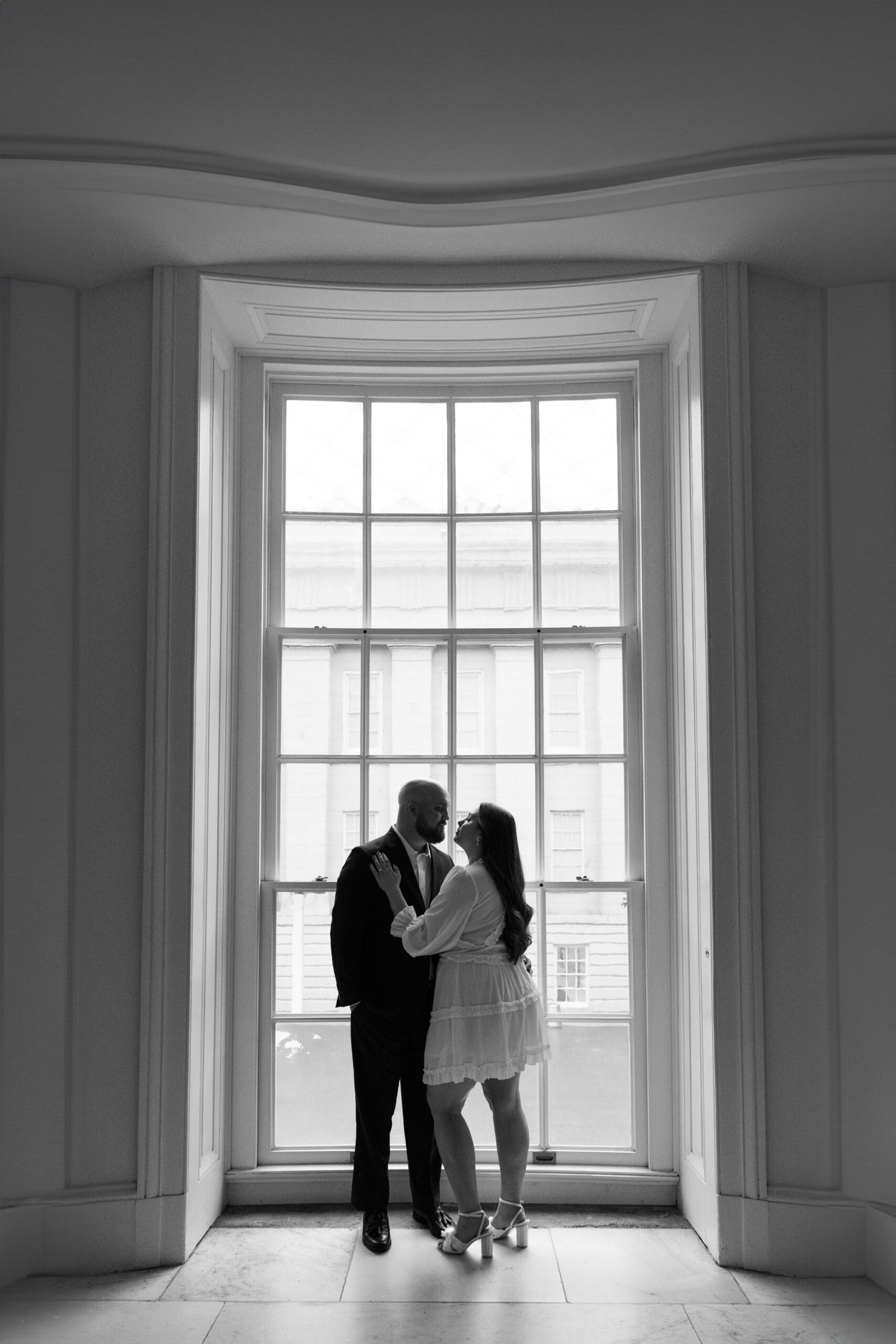 A couple embraces for their engagement photos at the National Portrait Gallery in Washington DC. They are standing in front of a large window.