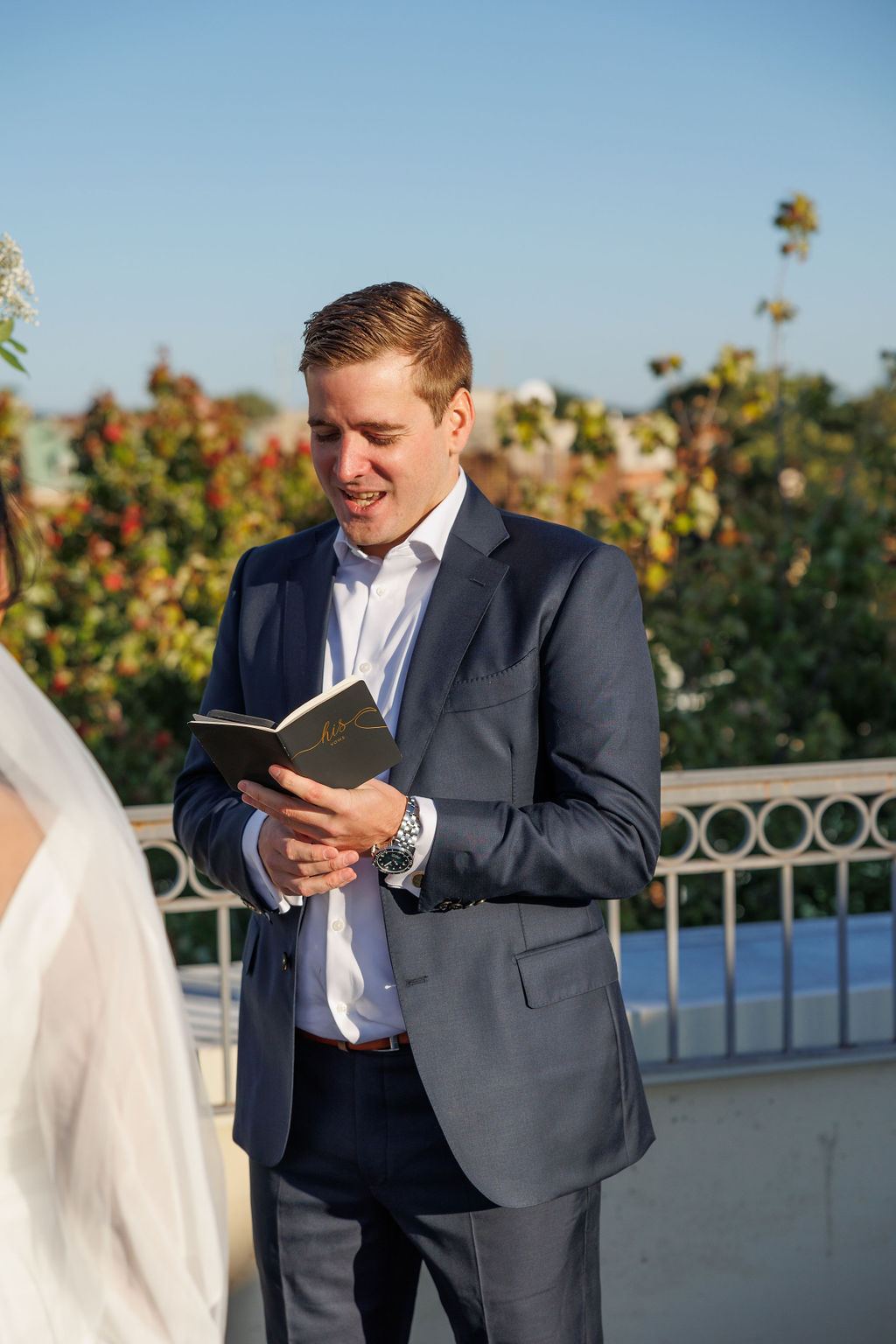A groom reads his vows from a leather book in a blue suit on a rooftop ceremony