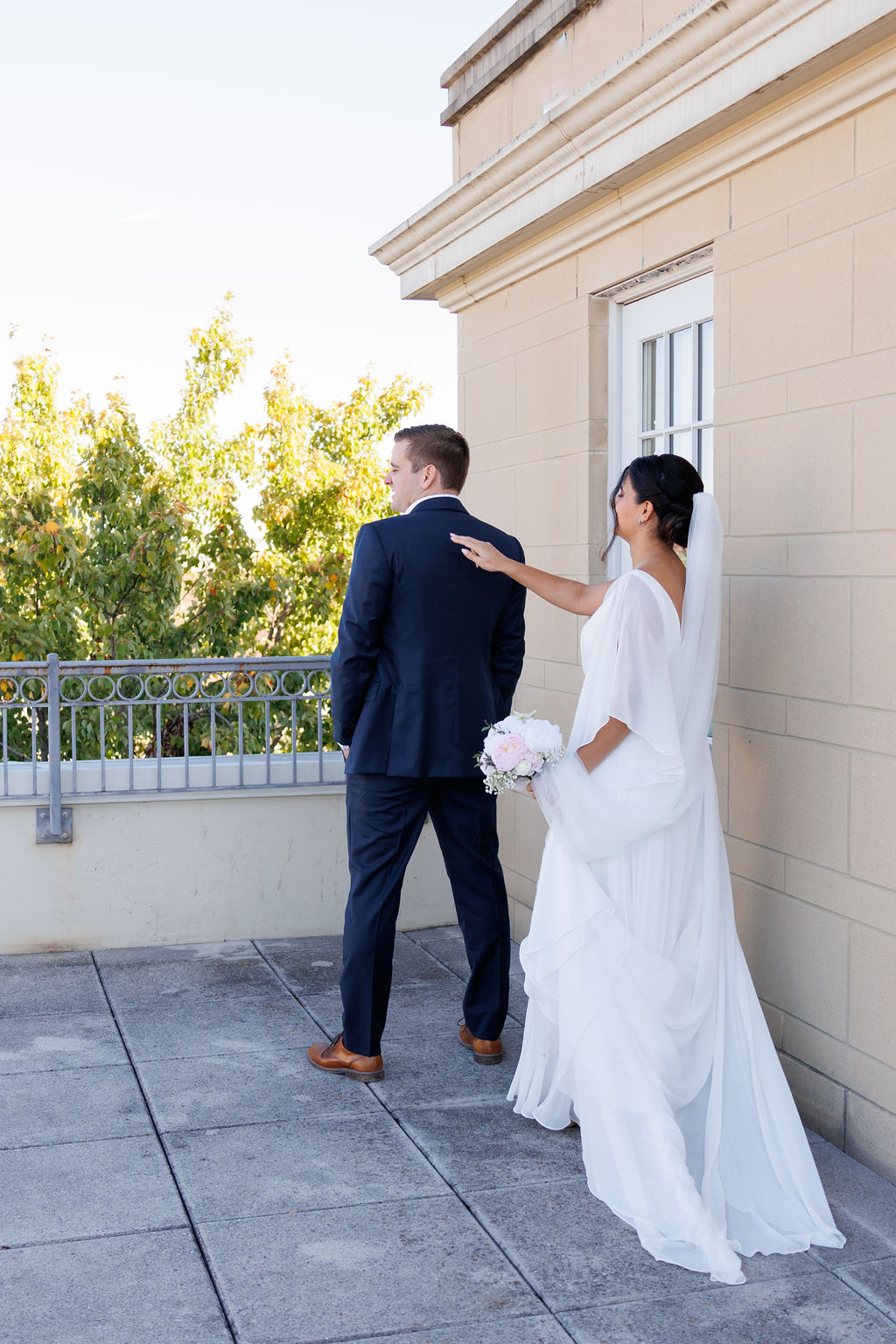 A groom waits for his bride to tap his shoulder on a rooftop balcony for their first look
