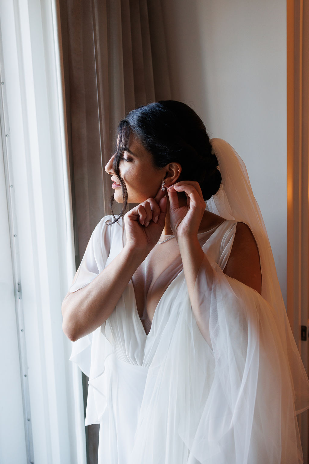 A bride stands in a window putting on her earrings