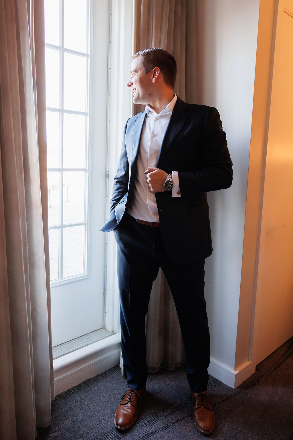 A groom stands in a window holding the lapel of his blue suit