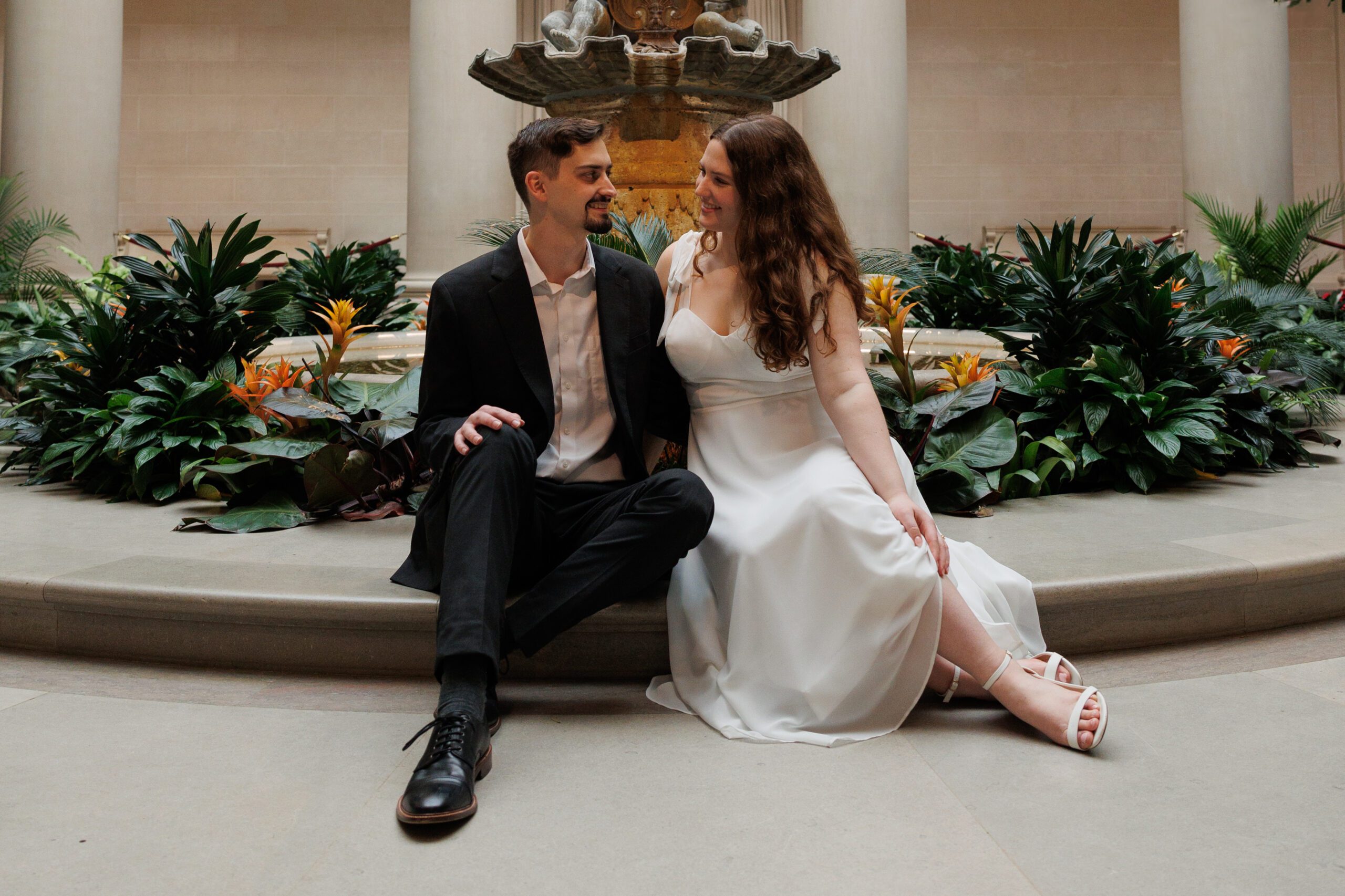 A newly engaged couple sits together in front of a fountain for their engagement photos in the west building in the national gallery of art